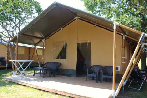 Tenda safari con veranda in legno coperta, sedie da esterno e alberi in un ambiente naturale.
