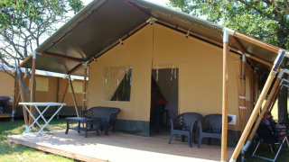 Safari tent with a covered wooden terrace, outdoor seating, and greenery in a relaxing camping area.