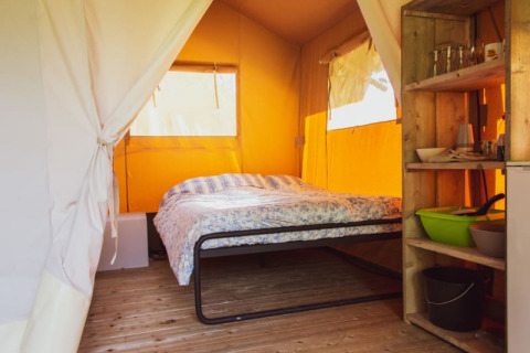 Interior view of a Safari tent with a double bed, open shelves, wooden floor and yellow canvas walls.