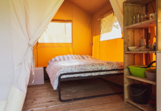 Interior view of a Safari tent with a double bed, open shelves, wooden floor and yellow canvas walls.
