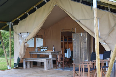 Interior view of a safari tent with wooden furniture, dining area, seating, and an open entrance to inside.