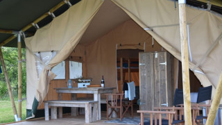 Innenbereich eines Safarizeltes mit Holztisch, Bänken und Stühlen auf einer Veranda im Freien.