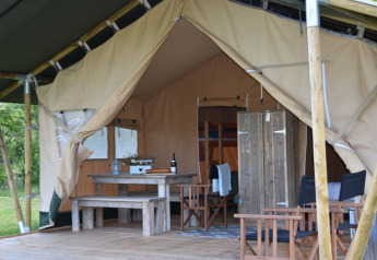 Interior view of a safari tent with wooden furniture, dining area, seating, and an open entrance to inside.