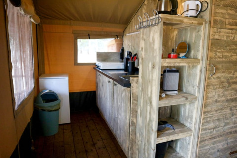 Kleine Küche in einem Safarizelt mit Holzregalen, Herd, Kühlschrank, Abfalleimer und Fenster.