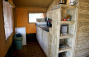 Angolo cucina in una tenda safari con mensole di legno, mini frigo, fornello e finestre luminose.