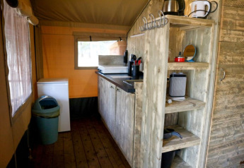 Kitchen area inside a safari tent featuring wooden shelves, stove, mini fridge, trash bin and windows.