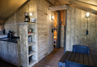 Vue intérieure d’une tente safari avec coin cuisine, table à manger, murs en bois et éclairage moderne.