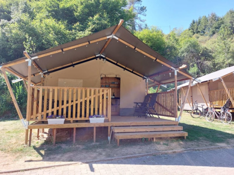 Safari tent with wooden porch in a green forest setting, bicycles and clear blue sky in the background.