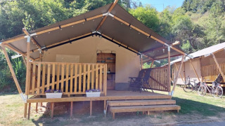Safari-Zelt mit Holzveranda und Fahrrädern davor, umgeben von Bäumen und sonnigem Himmel im Hintergrund.
