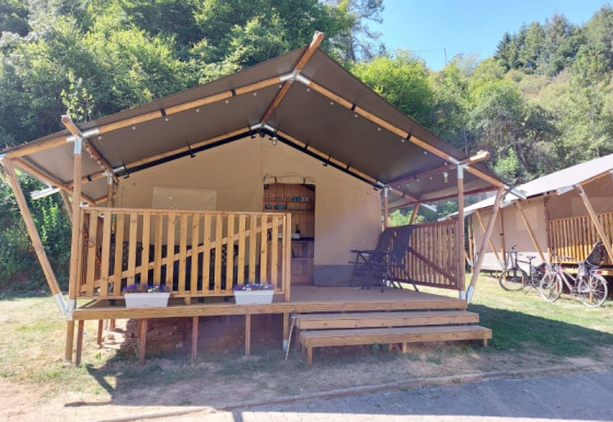 Safari tent with wooden porch in a green forest setting, bicycles and clear blue sky in the background.