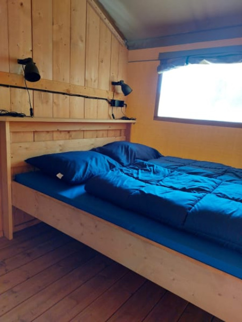 Innenaufnahme eines Holzbetts mit blauer Bettwäsche und Wandlampen im Safari-Zelt, Camping Drei Spatzen.