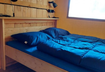 Vista interior de una cama de madera con ropa de cama azul y lámparas en una tienda safari, Camping Drei Spatzen.