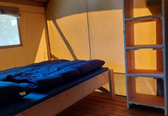 Vista interior de una tienda safari con cama, suelo de madera y estantería en Camping Drei Spatzen, Alemania.