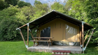 Safari tent on a grassy field, surrounded by lush trees and a bright blue sky with scattered clouds.