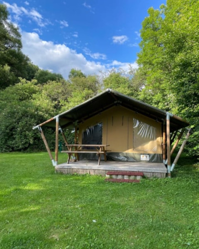 Safari tent on a grassy field, surrounded by lush trees and a bright blue sky with scattered clouds.