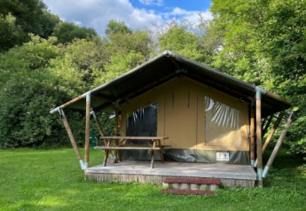 Safari tent on a grassy field, surrounded by lush trees and a bright blue sky with scattered clouds.