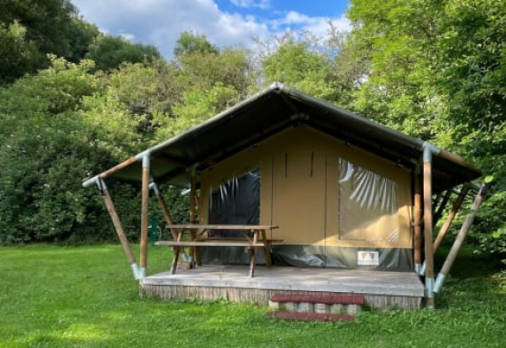 Safari tent on a grassy field, surrounded by lush trees and a bright blue sky with scattered clouds.
