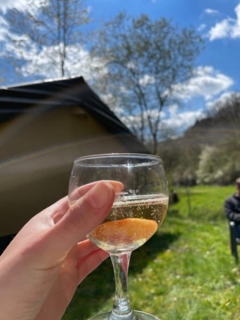 Hånd holder et glas mousserende vin foran en safaritelt på Camping Drei Spatzen under blå himmel i Tyskland.