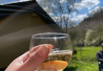 Hånd holder et glas mousserende vin foran en safaritelt på Camping Drei Spatzen under blå himmel i Tyskland.