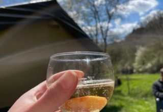 Hand met glas mousserende wijn voor een safaritent op Camping Drei Spatzen, Duitsland, onder een blauwe lucht.