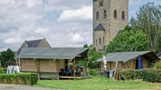 Safarizelt auf Campingplatz mit Steinkirche im Hintergrund und grüner Umgebung unter bewölktem Himmel.