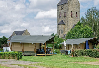 Tienda safari en camping con torre de iglesia de piedra al fondo y entorno verde bajo cielo nublado.