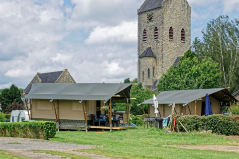 Tienda safari en camping con torre de iglesia de piedra al fondo y entorno verde bajo cielo nublado.