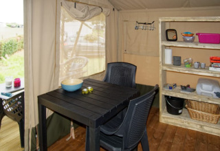 Vista interior de tienda safari en Camping La Dolce Vita, Países Bajos, con mesa, sillas y estantería.