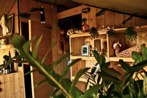 Interior view of a safari tent with wooden walls, jungle-themed plush toys, and green plants in the Netherlands.
