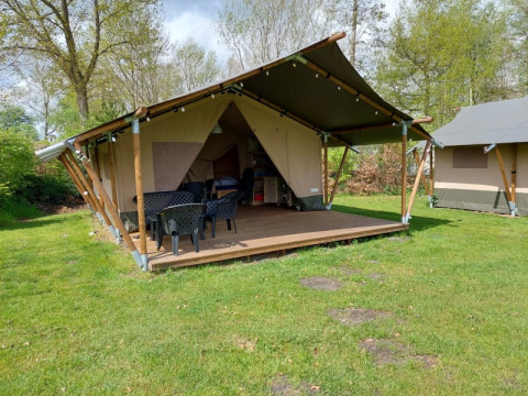 Safaritent met overdekte veranda, tuinstoelen en grasveld, omgeven door bomen op een zonnige dag.