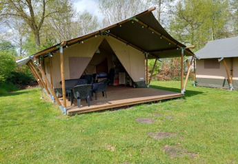 Safari tent with covered porch, outdoor seating, and green lawn surrounded by trees in daylight.