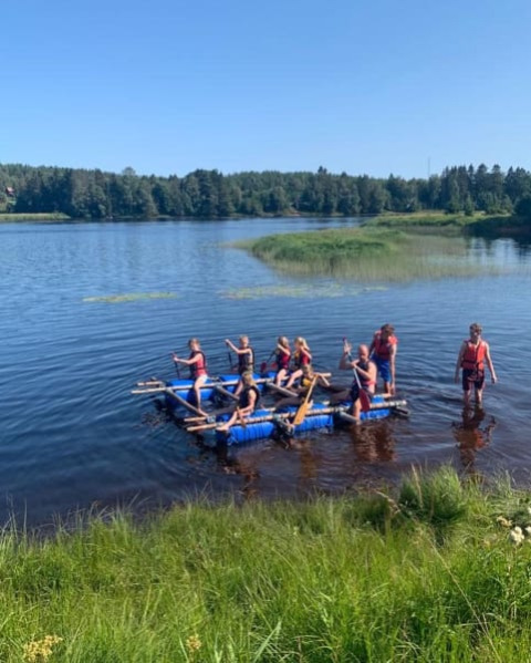Børn og voksne padler på en hjemmelavet tømmerflåde på en sø nær Safari telt på Camping Yttermalungs i Sverige.