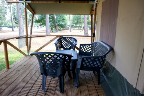 Terrasse en bois d'une tente safari avec chaises et table noires en plastique, entourée d’arbres en forêt.