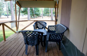 Terrasse en bois d'une tente safari avec chaises et table noires en plastique, entourée d’arbres en forêt.