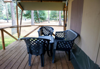 Terrasse en bois d'une tente safari avec chaises et table noires en plastique, entourée d’arbres en forêt.