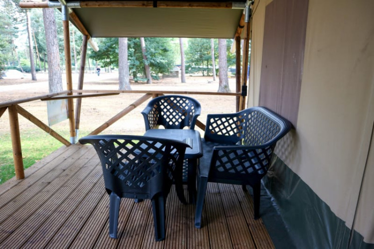 Wooden deck at a safari tent with black plastic chairs and table, surrounded by trees in a forest setting.