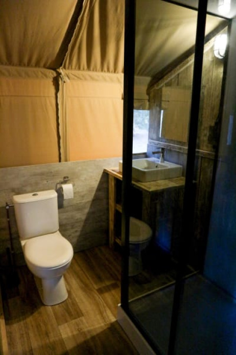 Salle de bain moderne dans une tente safari avec toilettes, lavabo, douche vitrée et murs en toile.
