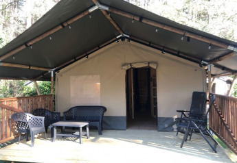 Safaritent + sanitair met overdekt houten terras en zitgedeelte buiten in een bosrijke omgeving.