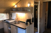 Zona de cocina en tienda safari con estufa, fregadero, nevera y baño en Camping Siësta en Bélgica.