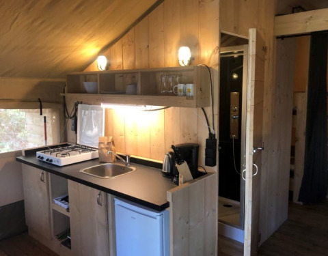 Zona de cocina en tienda safari con estufa, fregadero, nevera y baño en Camping Siësta en Bélgica.