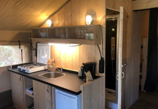 Zona de cocina en tienda safari con estufa, fregadero, nevera y baño en Camping Siësta en Bélgica.