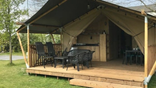 Outdoor photo of a safari tent with a wooden deck, seating area, and indoor kitchen plus bathroom facilities.