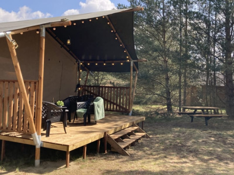 Tenda safari con veranda in legno, sedie, luci sospese e tavolo da picnic in area campeggio tra alberi.
