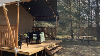 Tienda safari con terraza de madera, sillas, luces y mesa de picnic en un área de camping boscosa.
