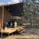 Safari telt med træterrasse, stole, lanterner og picnicbord i skovklædt campingområde under blå himmel.