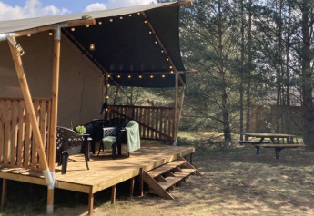 Tienda safari con terraza de madera, sillas, luces y mesa de picnic en un área de camping boscosa.