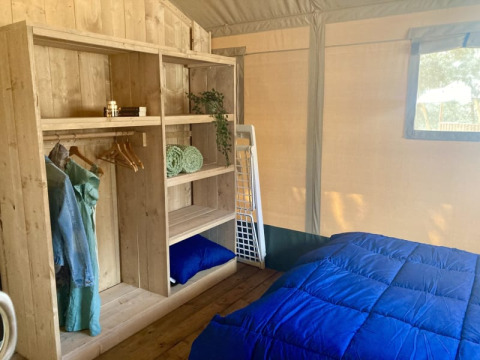 Innenraum eines Safari-Zeltes mit Holzregal, Kleiderbügeln, Kleidung und einem Bett mit blauer Decke.