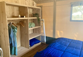 Innenraum eines Safari-Zeltes mit Holzregal, Kleiderbügeln, Kleidung und einem Bett mit blauer Decke.