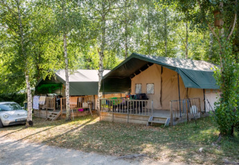 Twee safaritenten met groene daken staan tussen bomen, met een auto geparkeerd naast de camping.