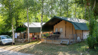Two safari tents with green roofs surrounded by leafy trees, with a car parked nearby on a campsite.